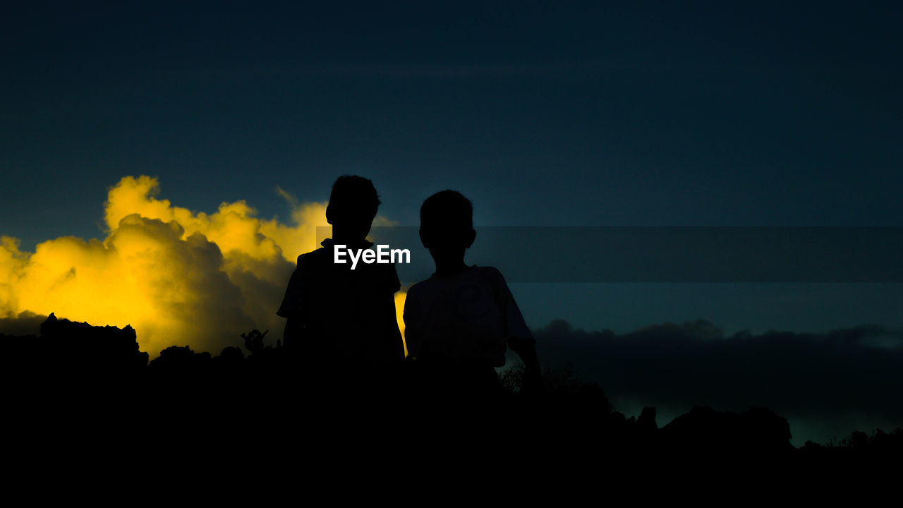 Silhouette siblings standing against sky at sunset