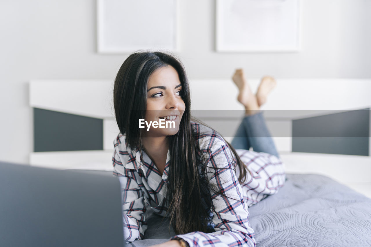 Smiling woman looking away while lying with laptop in bedroom