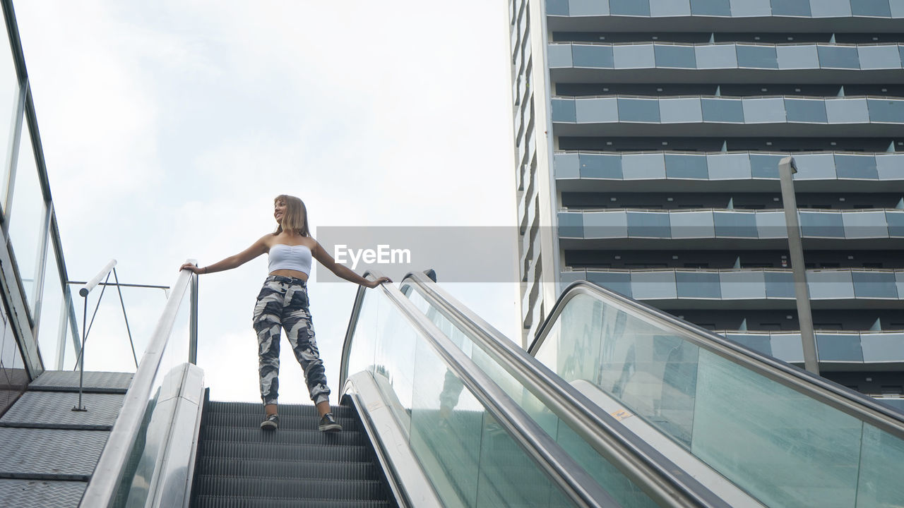 Low angle view of teenage girl standing on escalator against sky