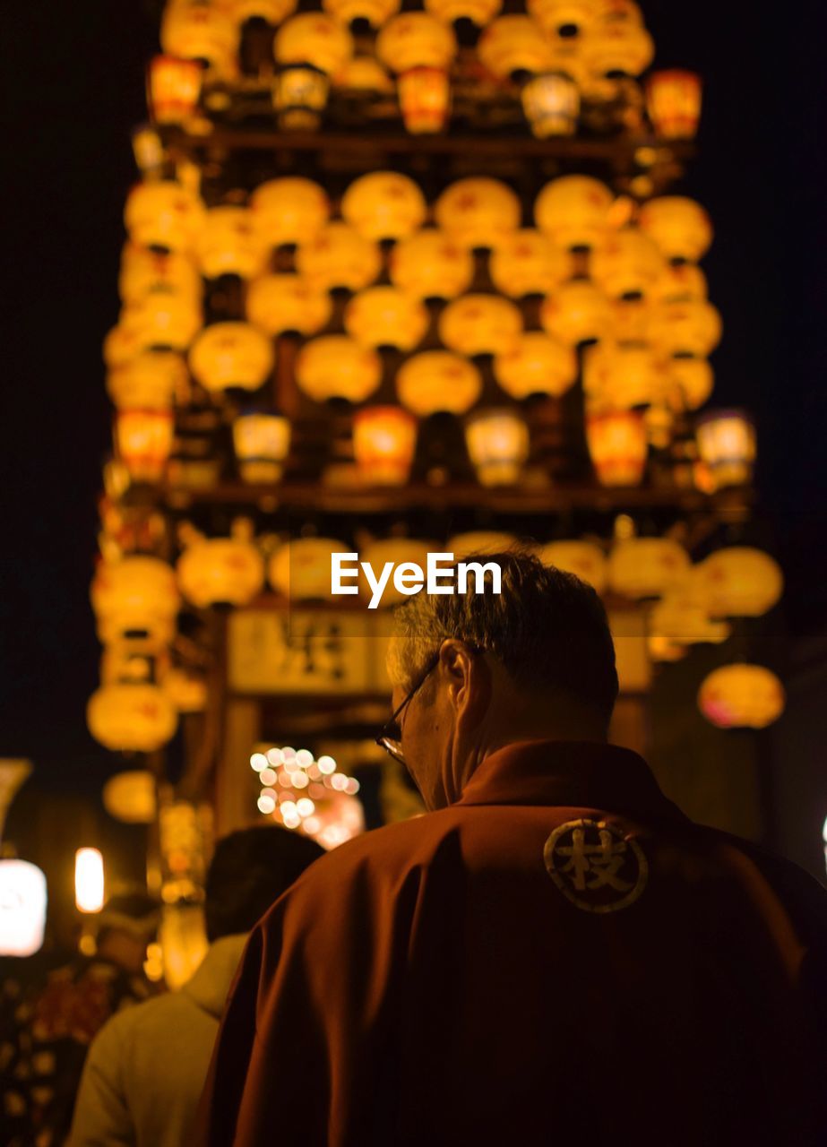 REAR VIEW OF MAN ON ILLUMINATED OUTSIDE TEMPLE AGAINST BUILDING