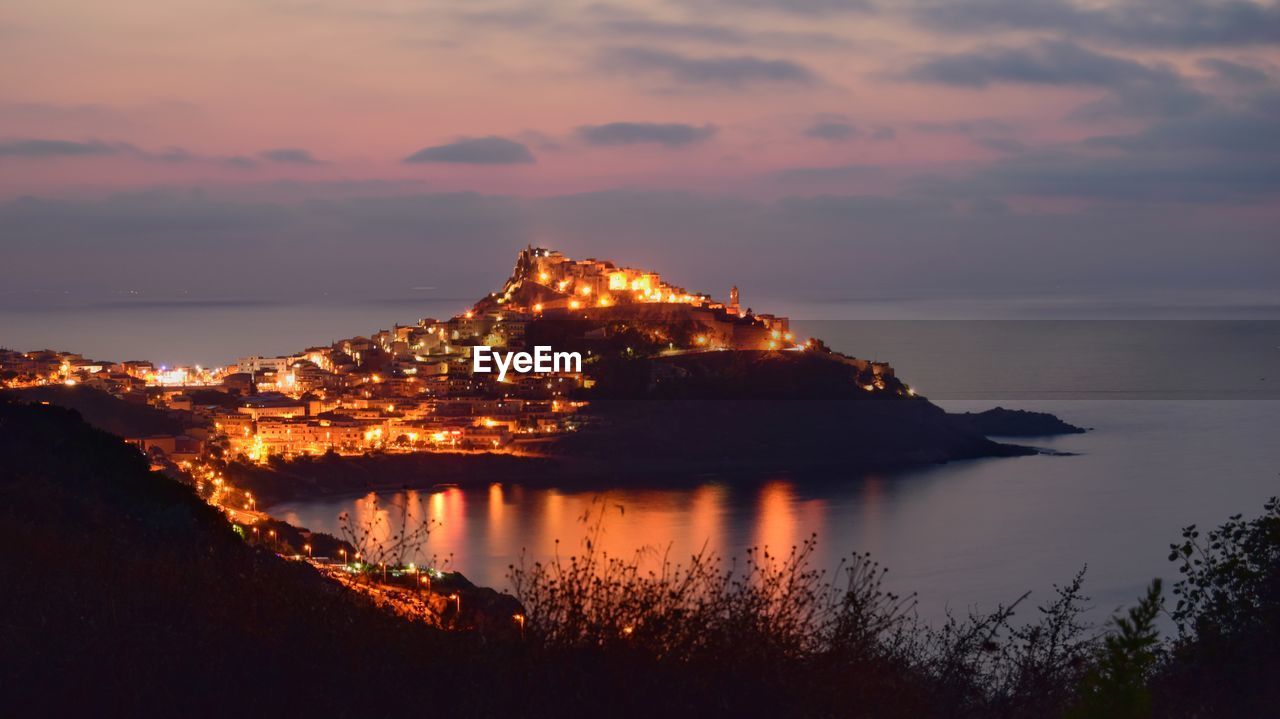 Illuminated city by sea against sky during sunset