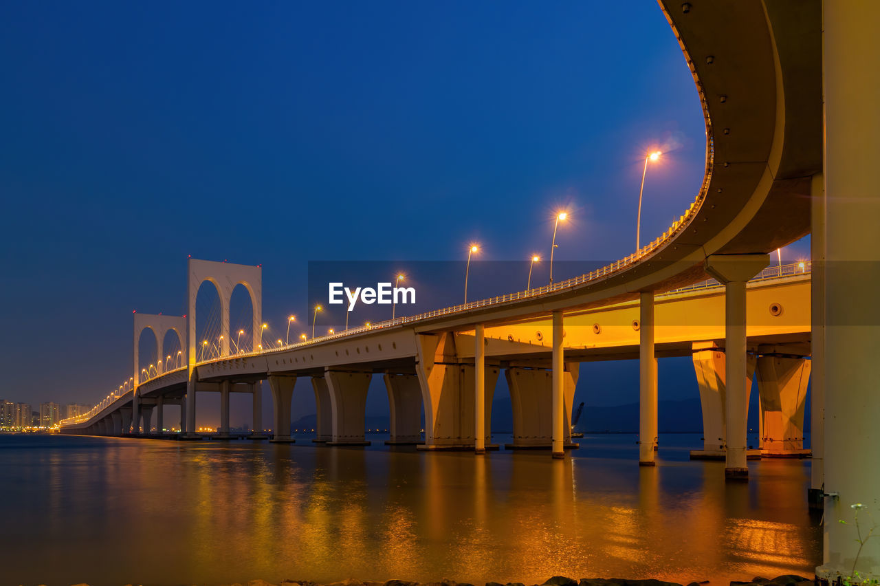 LOW ANGLE VIEW OF ILLUMINATED BRIDGE OVER RIVER AGAINST SKY AT NIGHT