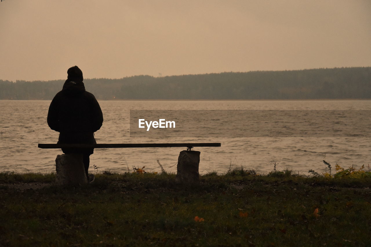 REAR VIEW OF MAN SITTING ON SHORE AT SUNSET