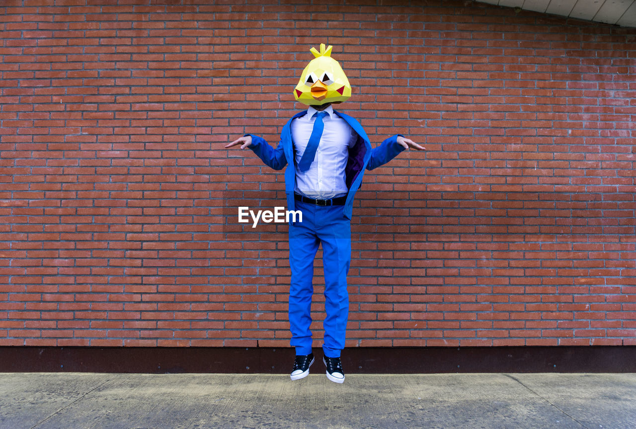 Man wearing vibrant blue suit and bird mask pretending to fly in front of brick wall