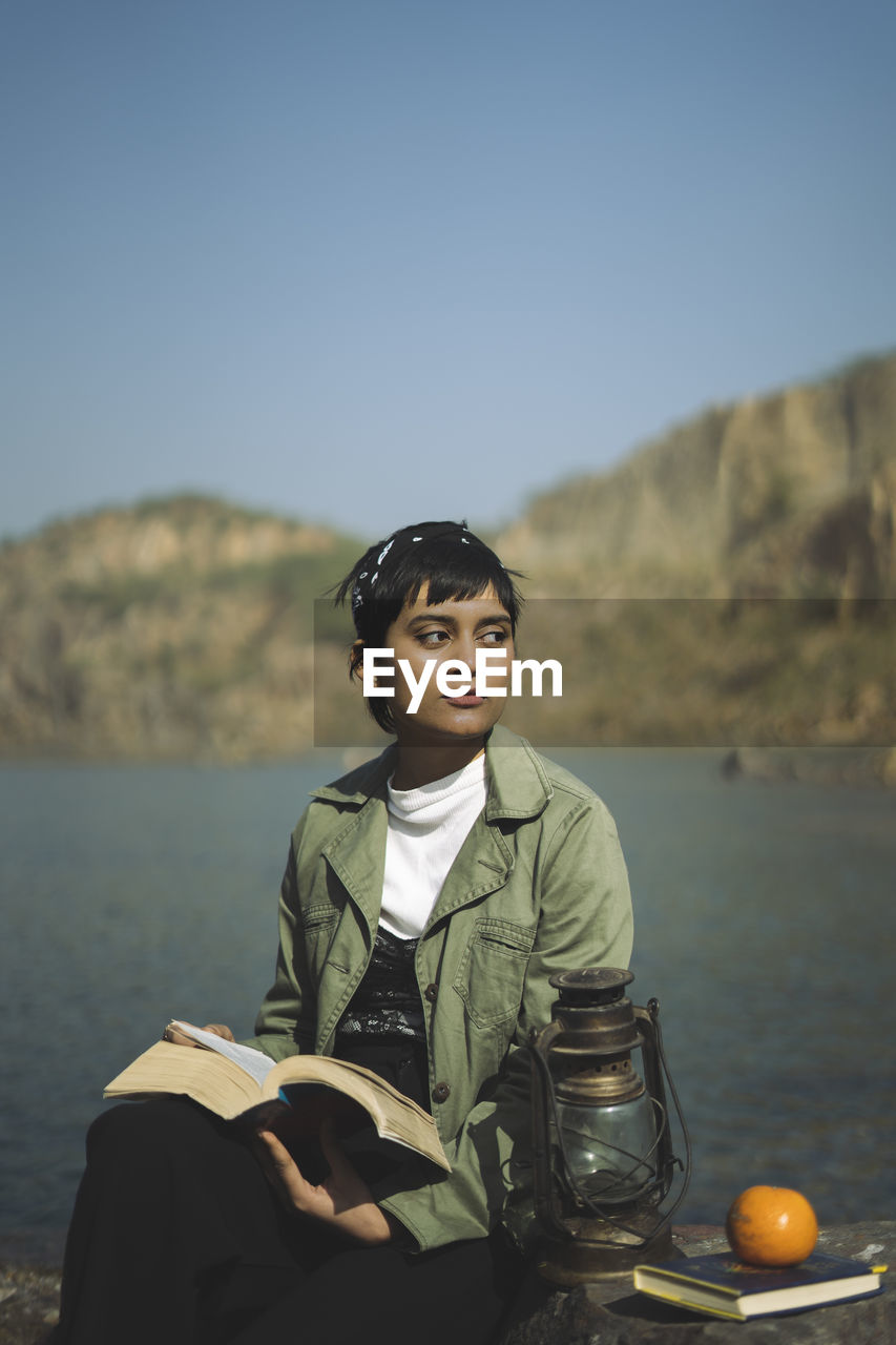 Young woman looking away while holding book sitting by lake