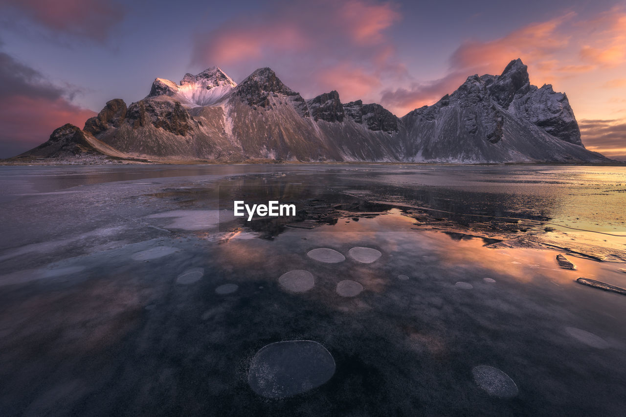 Spectacular nordic scenery of calm frozen lake near rocky vestrahorn mountain with snowy peaks during colorful sunset at stockness beach, iceland