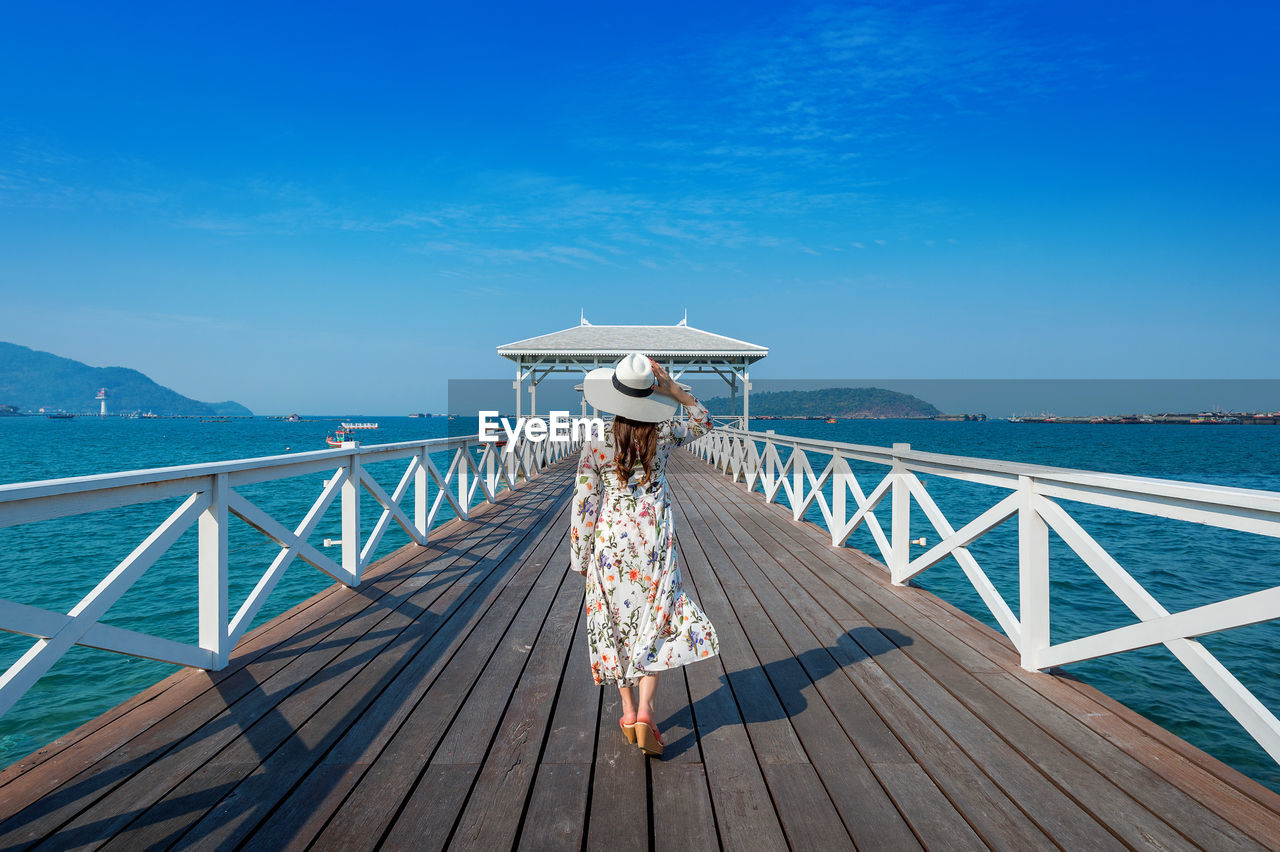 Rear view of woman walking on pier over sea against blue sky