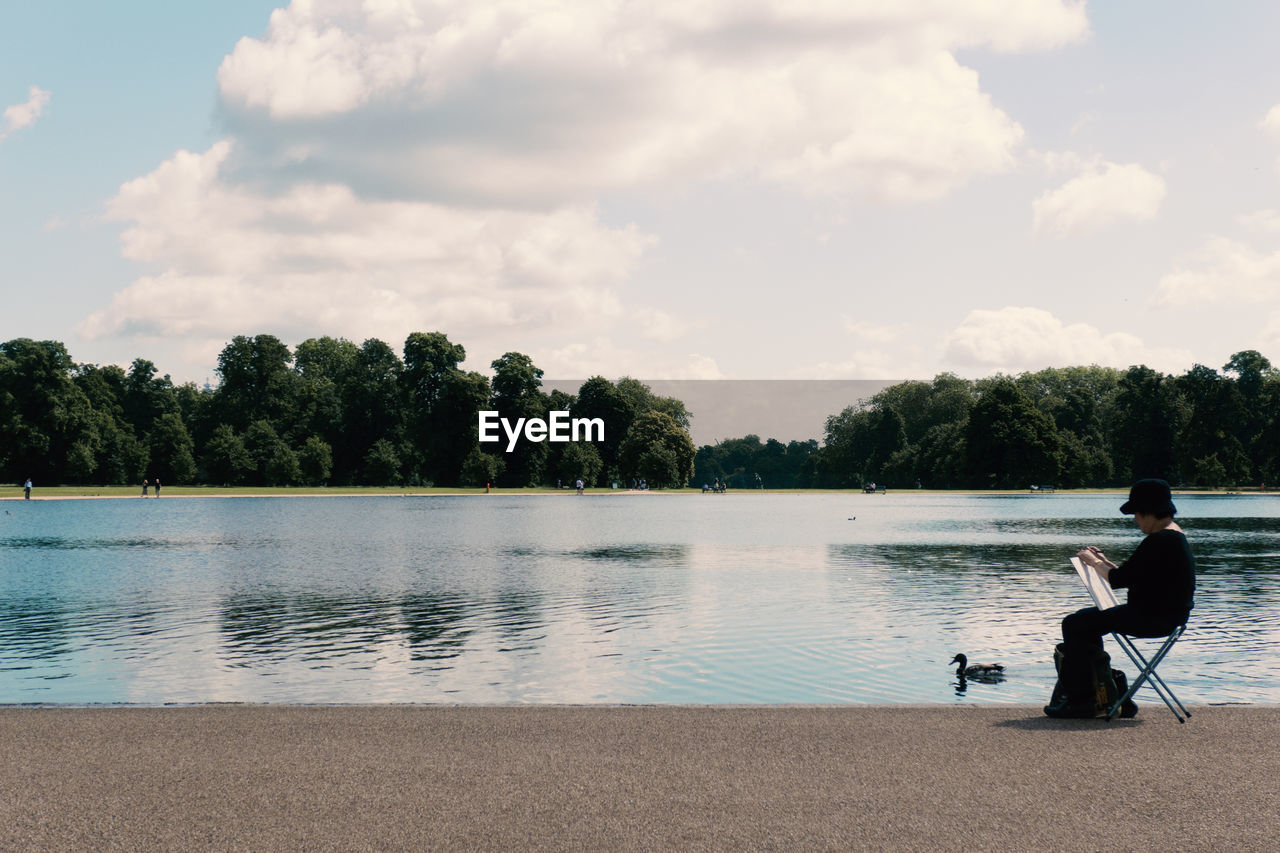 Side view of person holding paper while sitting by bird in lake against sky