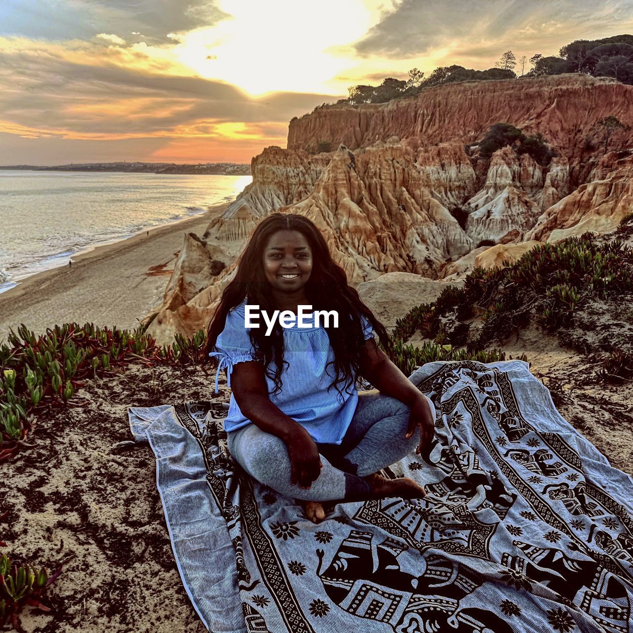 PORTRAIT OF SMILING YOUNG WOMAN SITTING ON ROCK AGAINST SUNSET