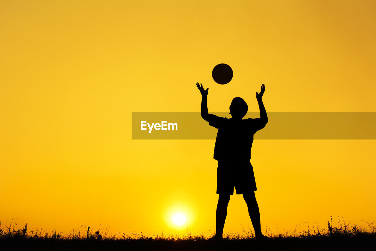 Silhouette boy playing with ball while standing on field against orange sky