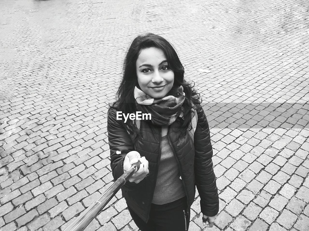 High angle portrait of woman holding monopod while standing on cobbled footpath