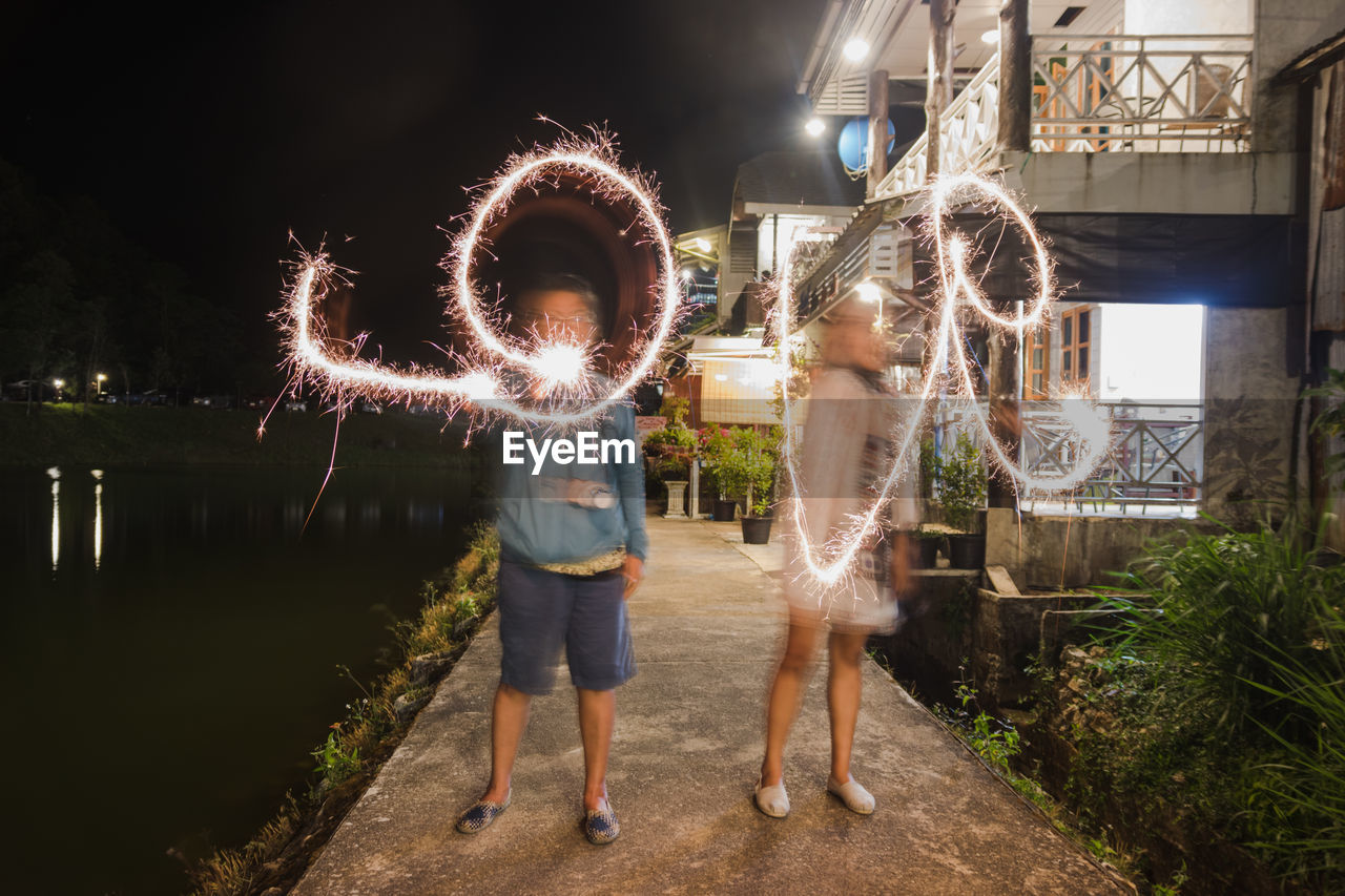 Friends making love text with sparkler while standing by lake against sky at night