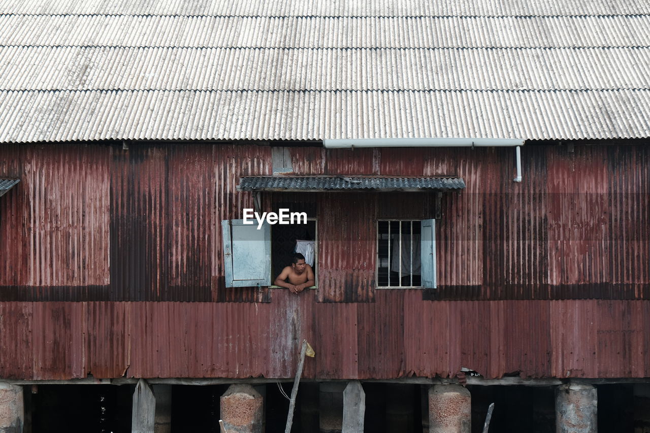 Shirtless man looking through window of house