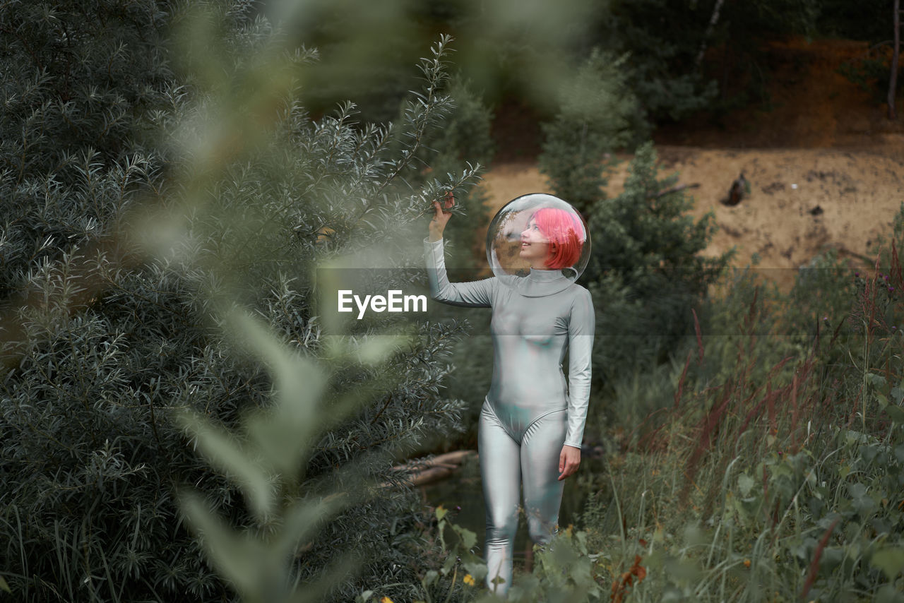 Happy young red haired female astronaut in silver suit and glass sphere helmet looking away while walking in field near forest in summer day