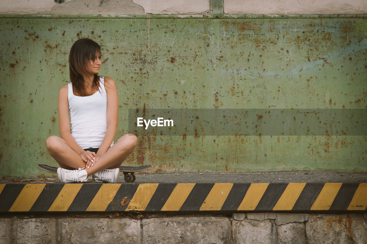 Young woman looking away while sitting on skateboard against wall