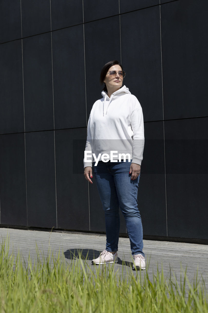 Full length sportive mature brunette woman in white hoody, blue jeans and sunglasses looks away