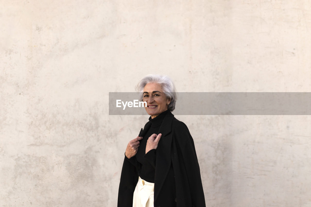 Smiling mature woman wearing jacket by wall