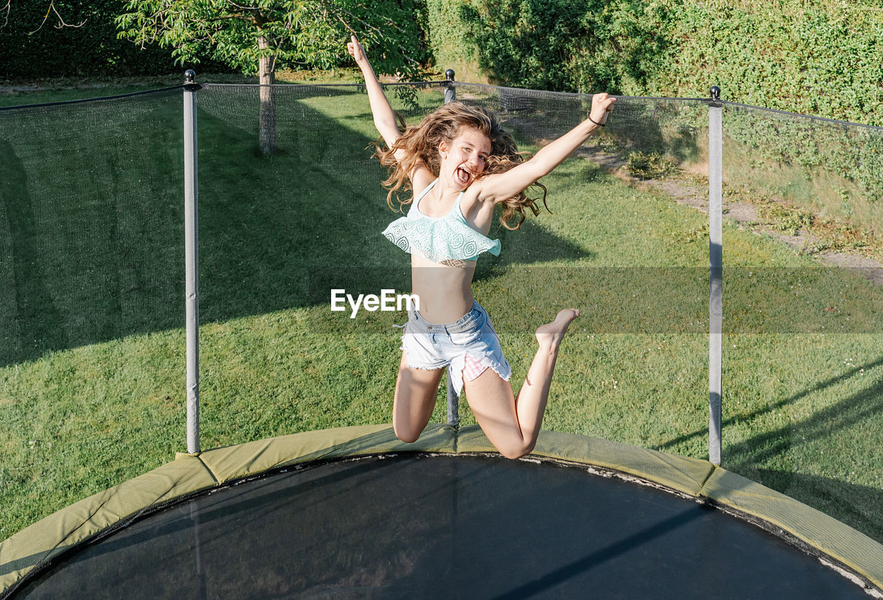 Horizontal photo of a smiling young brunette teenager jumping on a trampoline with net around on the green yard outdoors. the girl wears short jeans and a top and looks fun, active