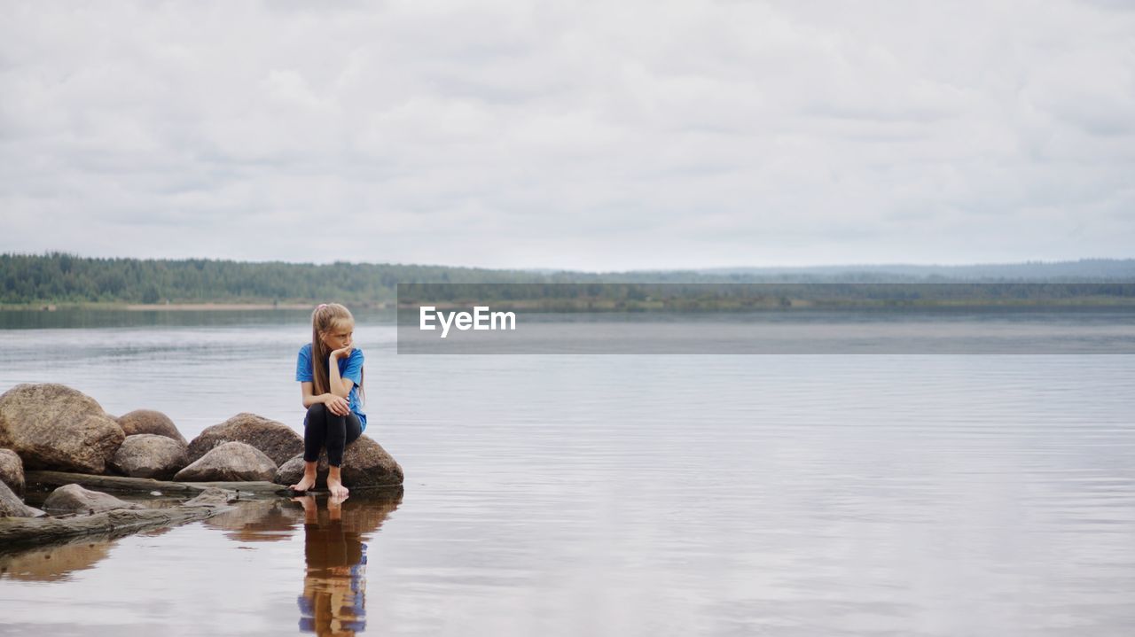 Teenage girl sitting on rock in lake against cloudy sky