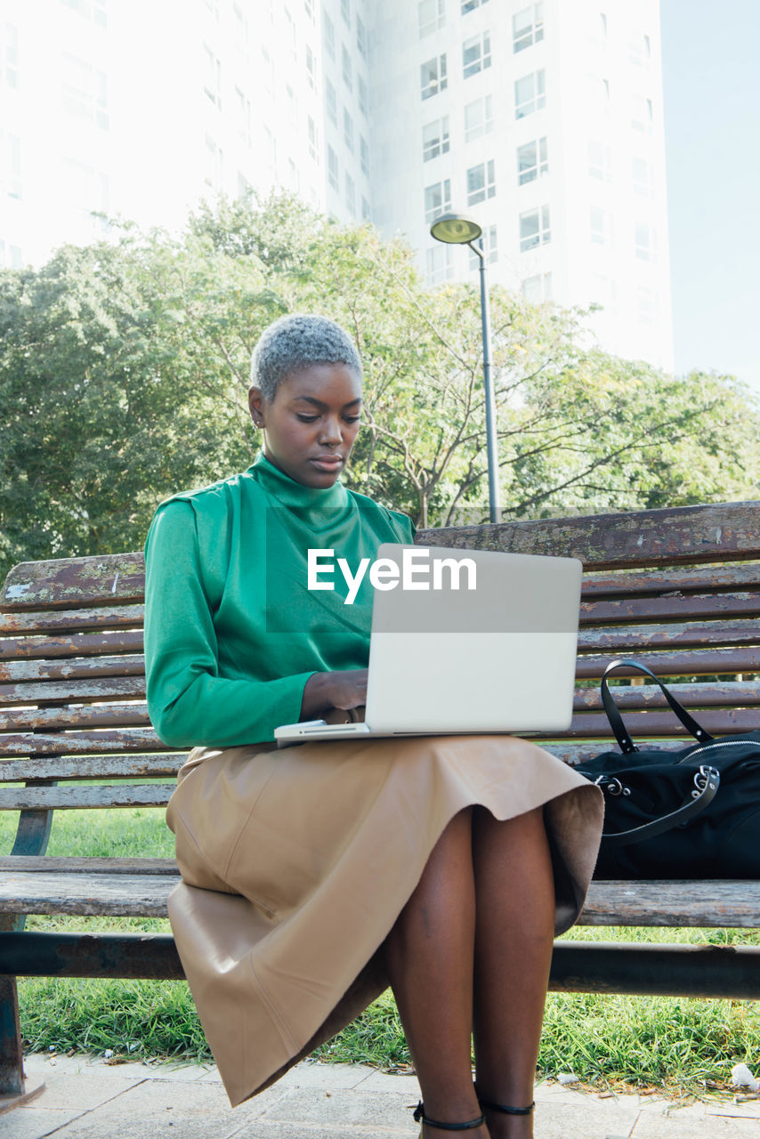 Concentrated young african american female freelancer with short dyed hair in stylish clothes sitting on wooden bench in park and working remotely on laptop