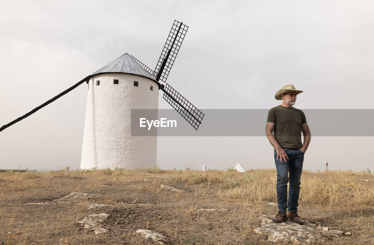 Man with cowboy hat standing by spanish traditional white windmills against sky