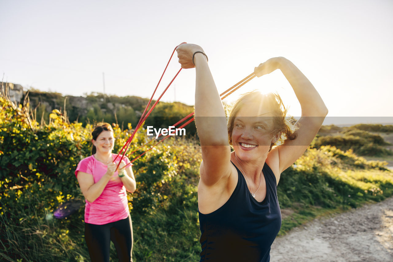 Smiling woman holding resistance band while exercising with woman at beach