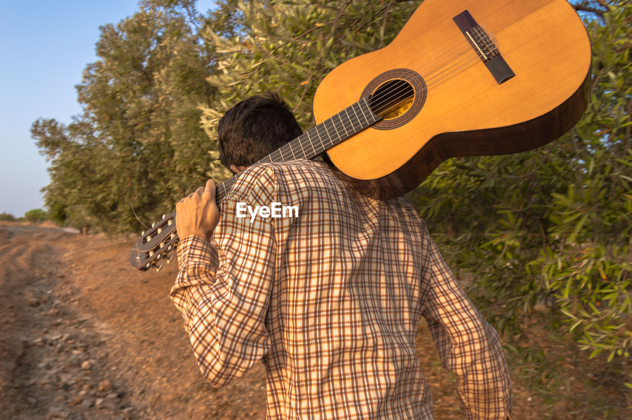 Rear view of man with acoustic guitar walking by trees on field 
