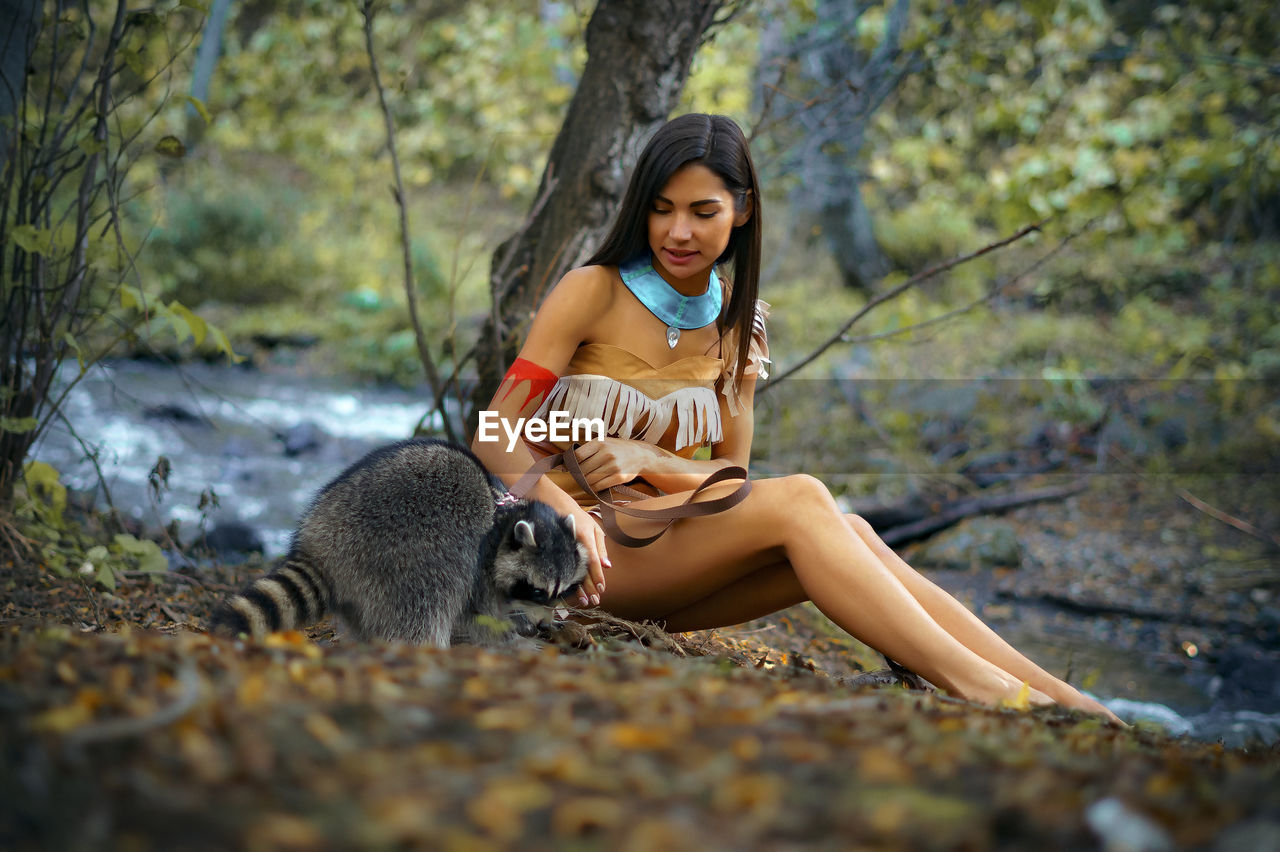 Smiling young woman in traditional clothing sitting by raccoon in forest