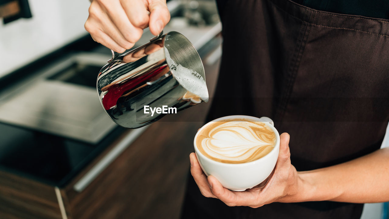 Crop image of a young male barista pouring hot milk into espresso black coffee for making latte art.