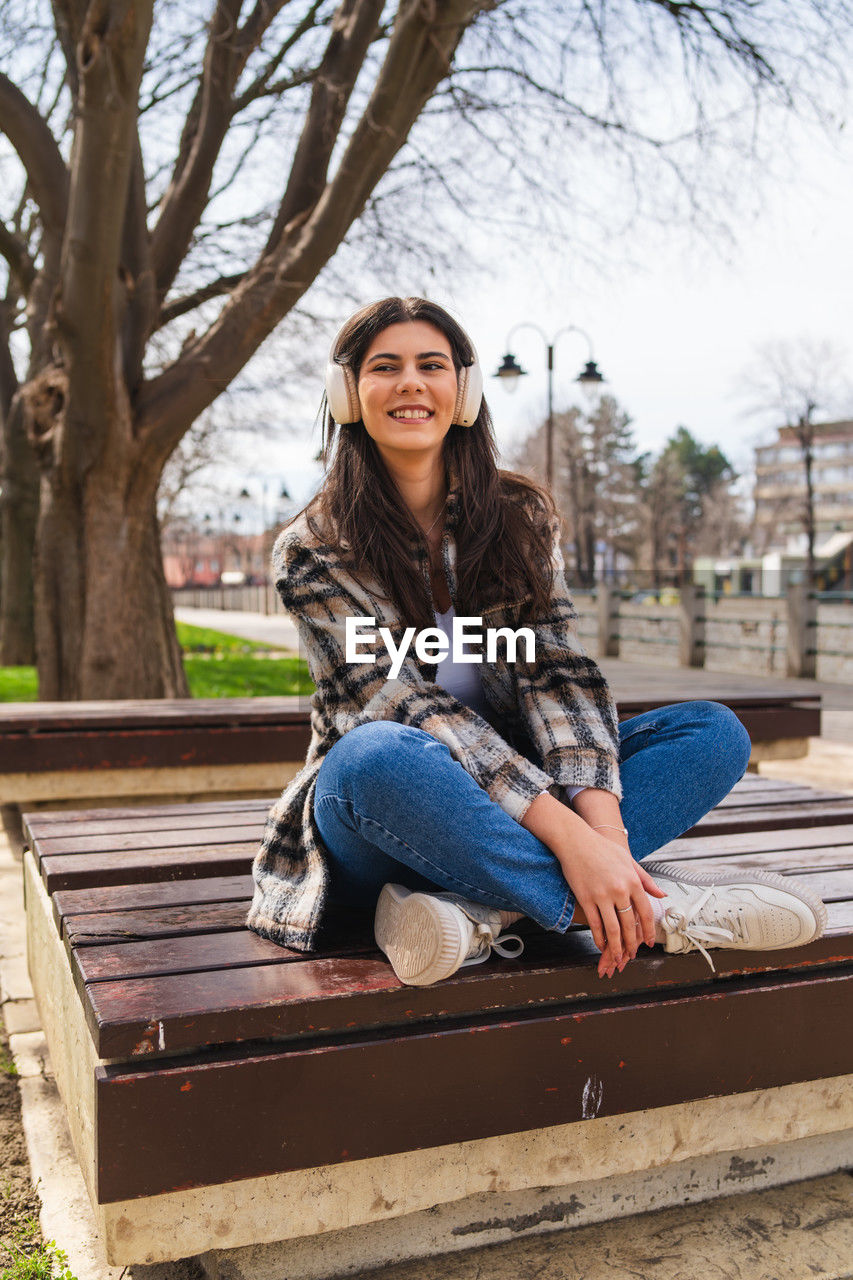 one person, sitting, long hair, smiling, women, portrait, young adult, casual clothing, adult, happiness, hairstyle, looking at camera, emotion, bench, full length, spring, brown hair, tree, leisure activity, lifestyles, jeans, nature, clothing, photo shoot, front view, architecture, day, park, relaxation, person, park bench, teeth, cheerful, park - man made space, outdoors, enjoyment, smile, city, plant, seat, female, teenager, fashion, child