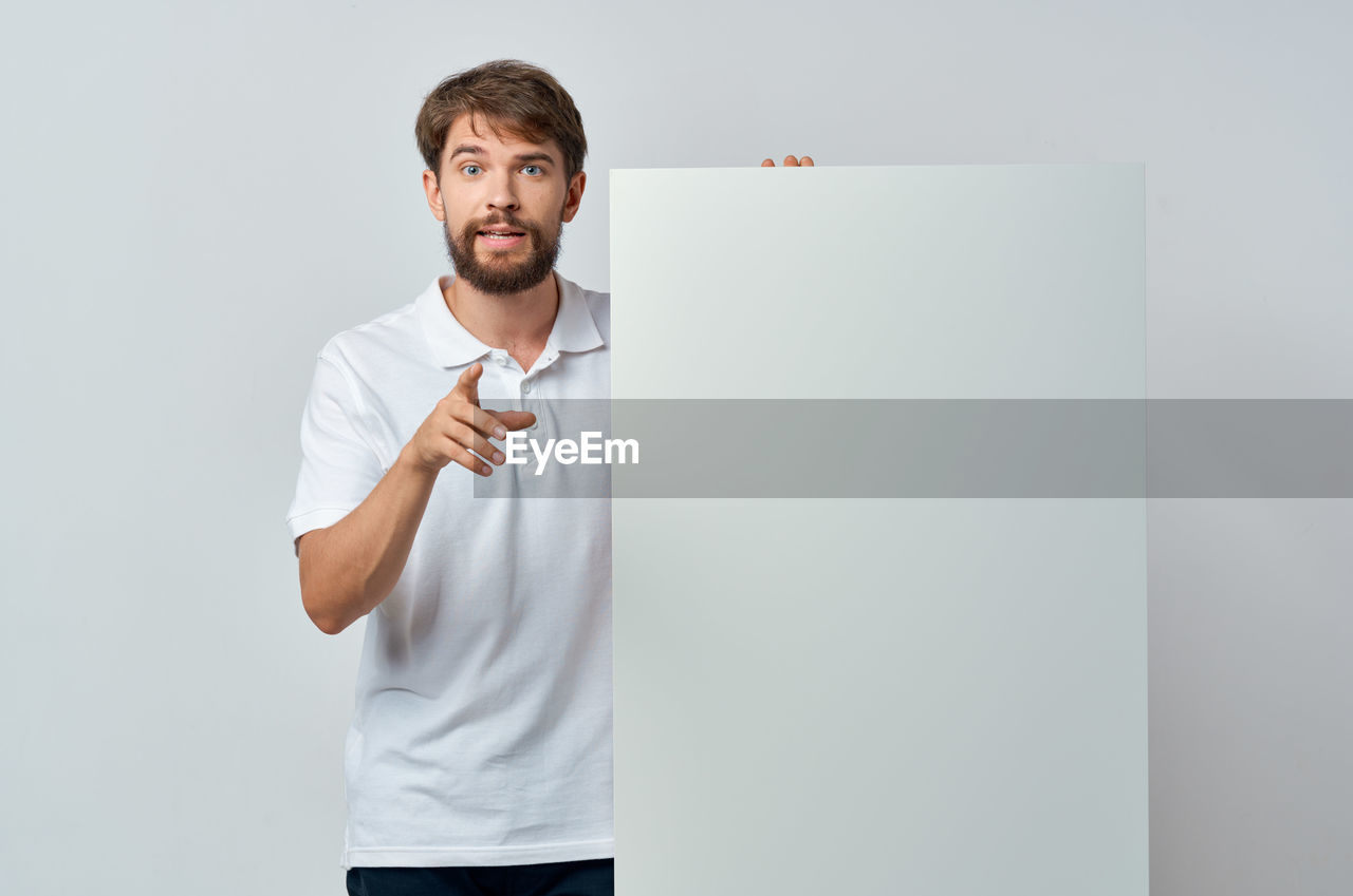 one person, adult, men, indoors, standing, beard, t-shirt, copy space, facial hair, casual clothing, young adult, white, studio shot, front view, portrait, clothing, whiteboard, waist up, business, looking, person, creativity, holding, sleeve, businessman, board, looking at camera, emotion