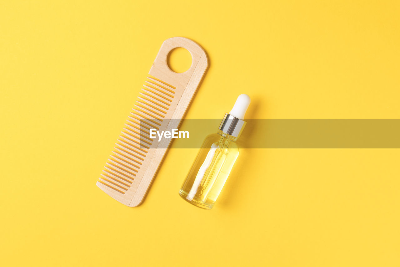 Natural hair care oil and wooden comb on yellow background, top view.