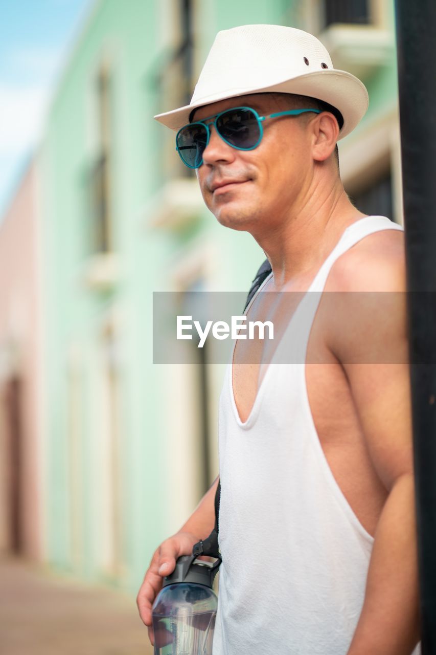 adult, one person, sunglasses, architecture, fashion, hat, glasses, clothing, lifestyles, spring, men, standing, portrait, sports, day, goggles, person, fashion accessory, city, outdoors, waist up, leisure activity, summer, young adult, focus on foreground, smiling, happiness, casual clothing, built structure, building exterior, exercising, nature, water, cap, baseball cap, eyewear, blue, relaxation