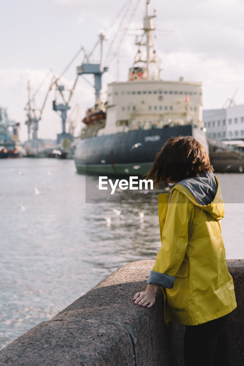Woman traveler in raincoat admiring the icebreaker and cranes at shipyard. selective focus on girl
