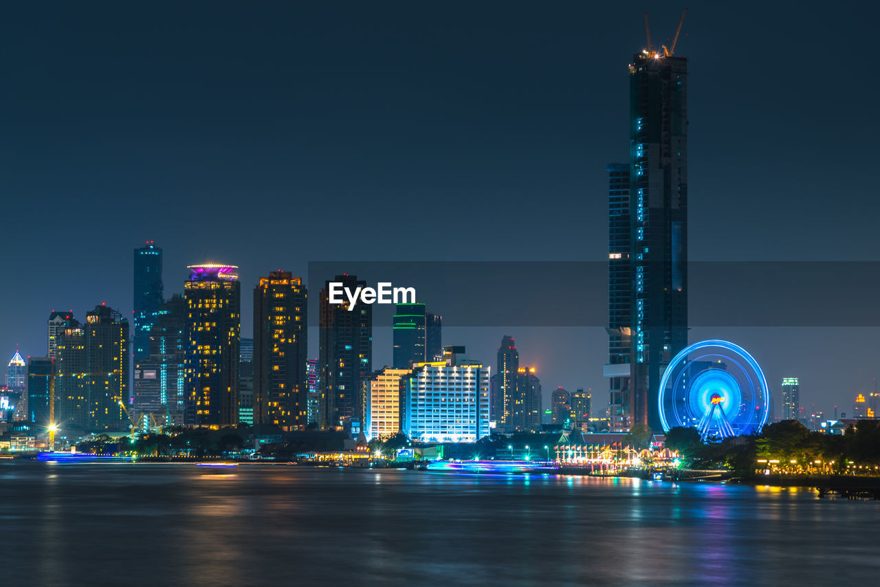 Illuminated buildings in front of chao phraya river against clear sky at night