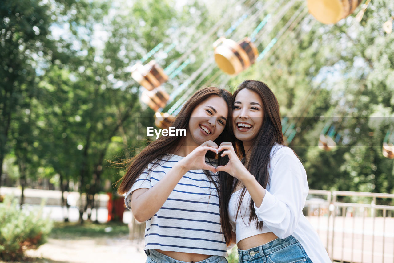 Young women with long hair friends having fun showing heart with theit hands at amusement park