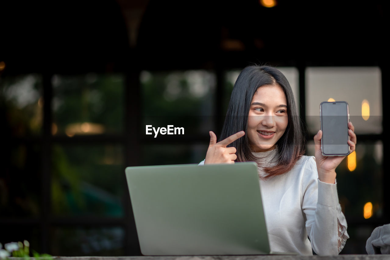 PORTRAIT OF SMILING YOUNG WOMAN USING MOBILE PHONE AT CAMERA