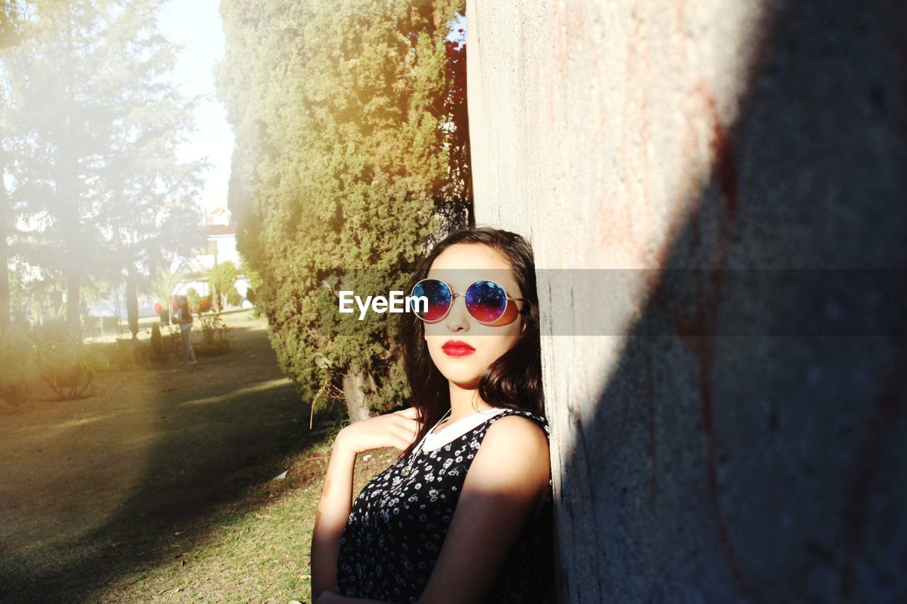 PORTRAIT OF YOUNG WOMAN WEARING SUNGLASSES STANDING AGAINST TREE