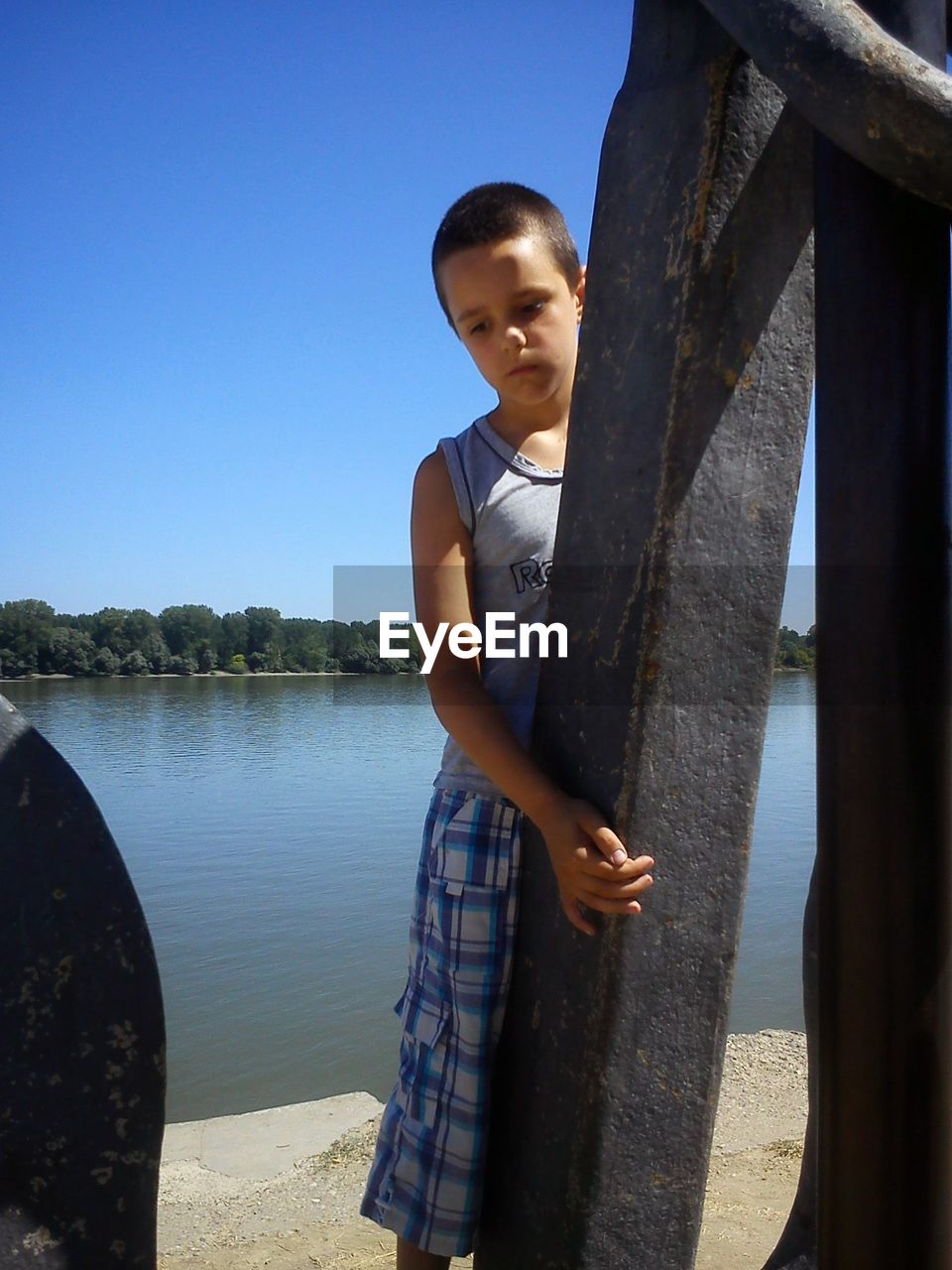 Boy standing by metallic anchor at lakeshore against sky