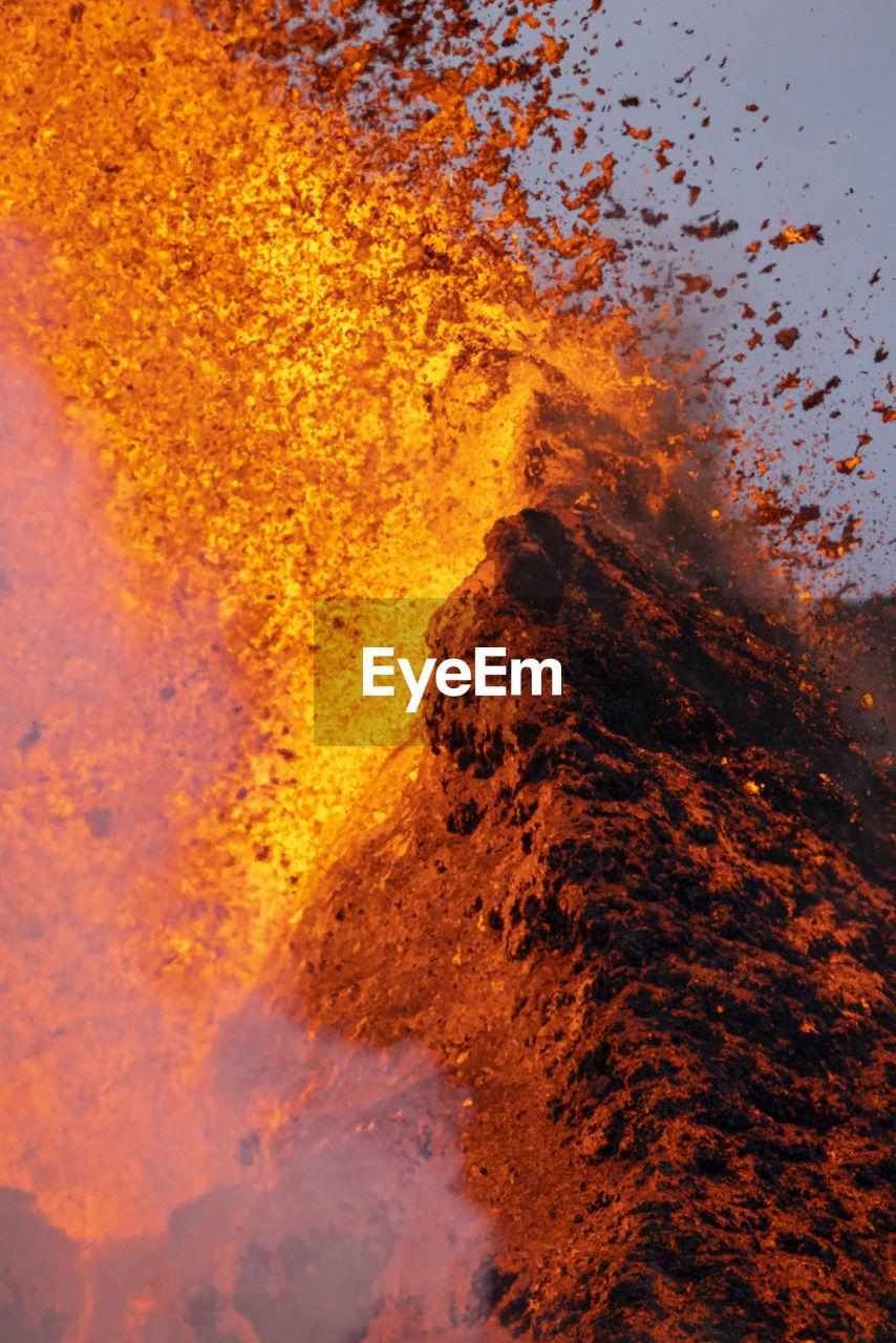 Splashes of hot orange lava erupting from volcanic mountain peak surrounded with smoke in iceland