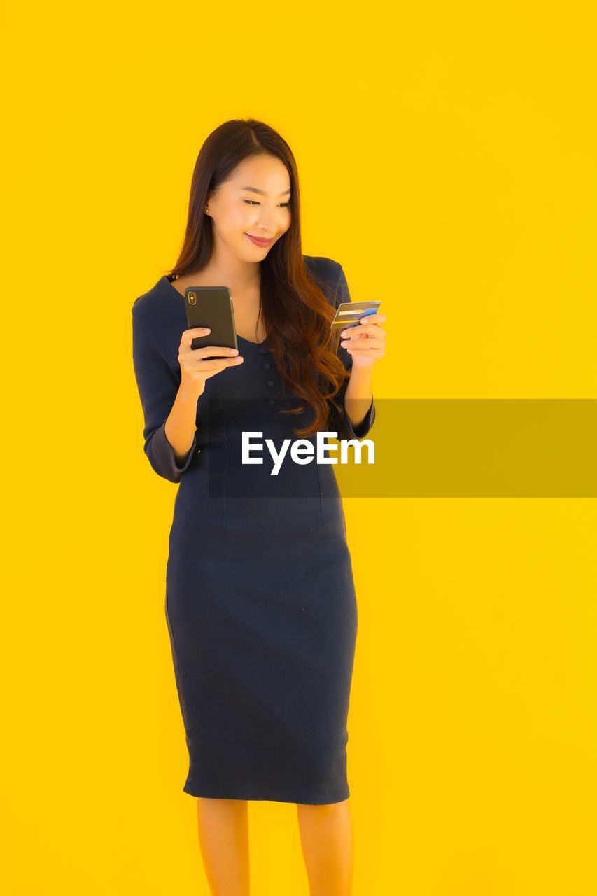 SMILING YOUNG WOMAN USING PHONE AGAINST YELLOW BACKGROUND