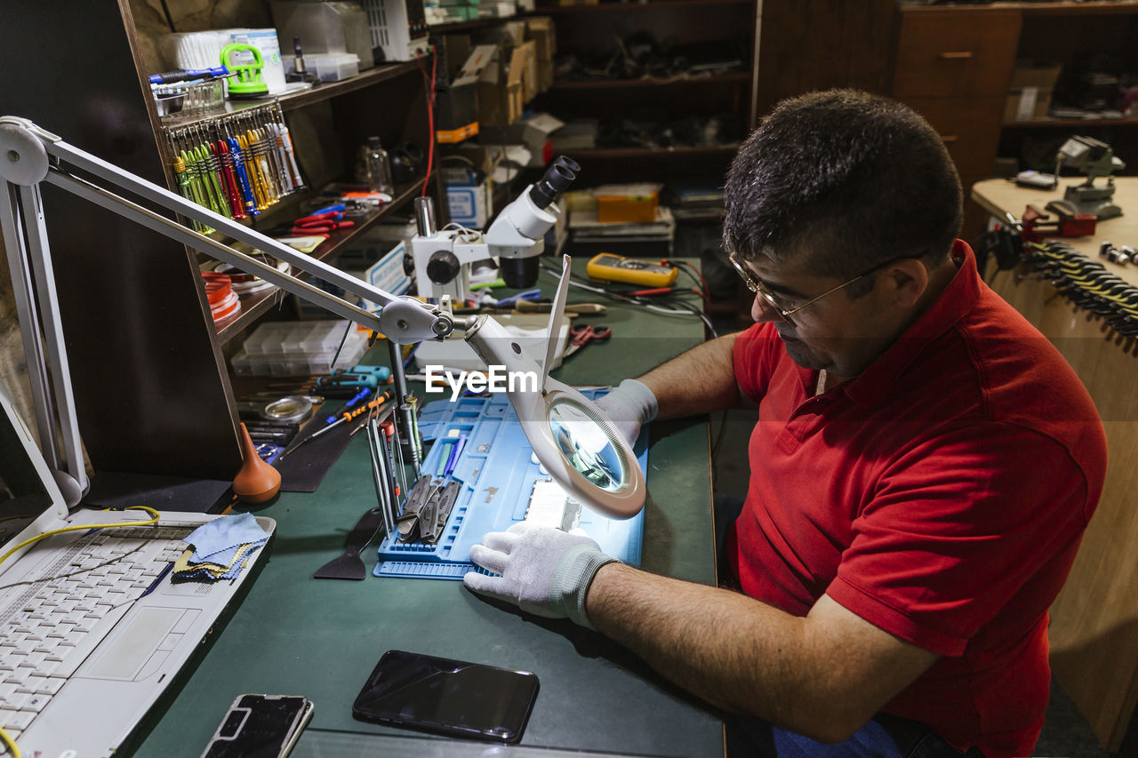 Worker looking at broken smart phone through magnifying glass in repair shop