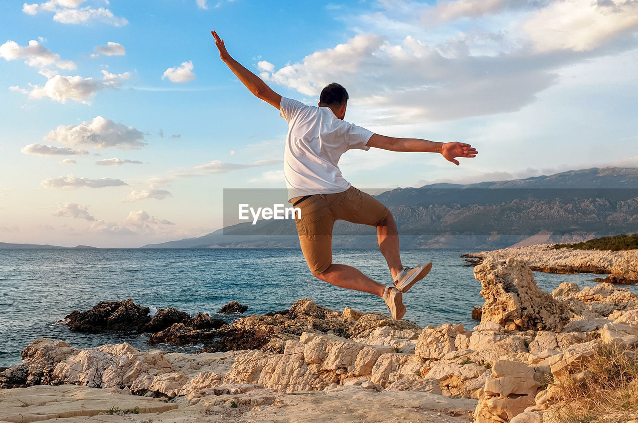 FULL LENGTH OF YOUNG MAN JUMPING ON ROCK AGAINST SEA
