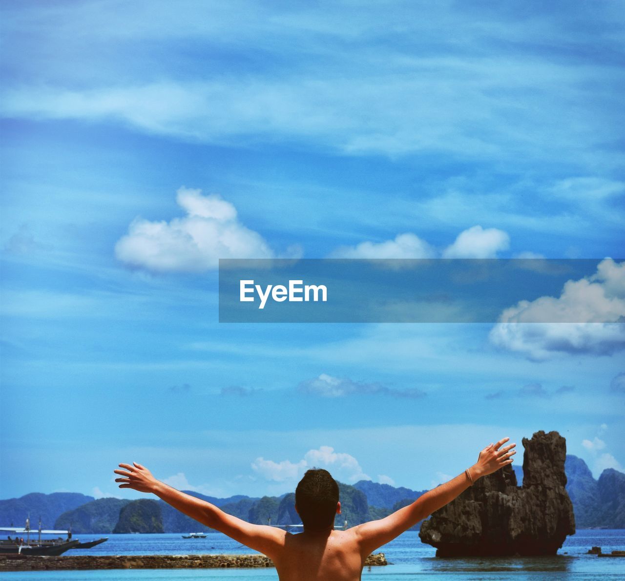 Rear view of shirtless man with arms outstretched against sky