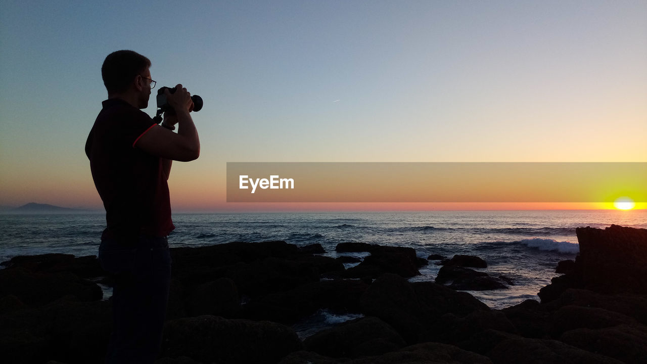 Side view of man photographing sea through digital camera at sunset