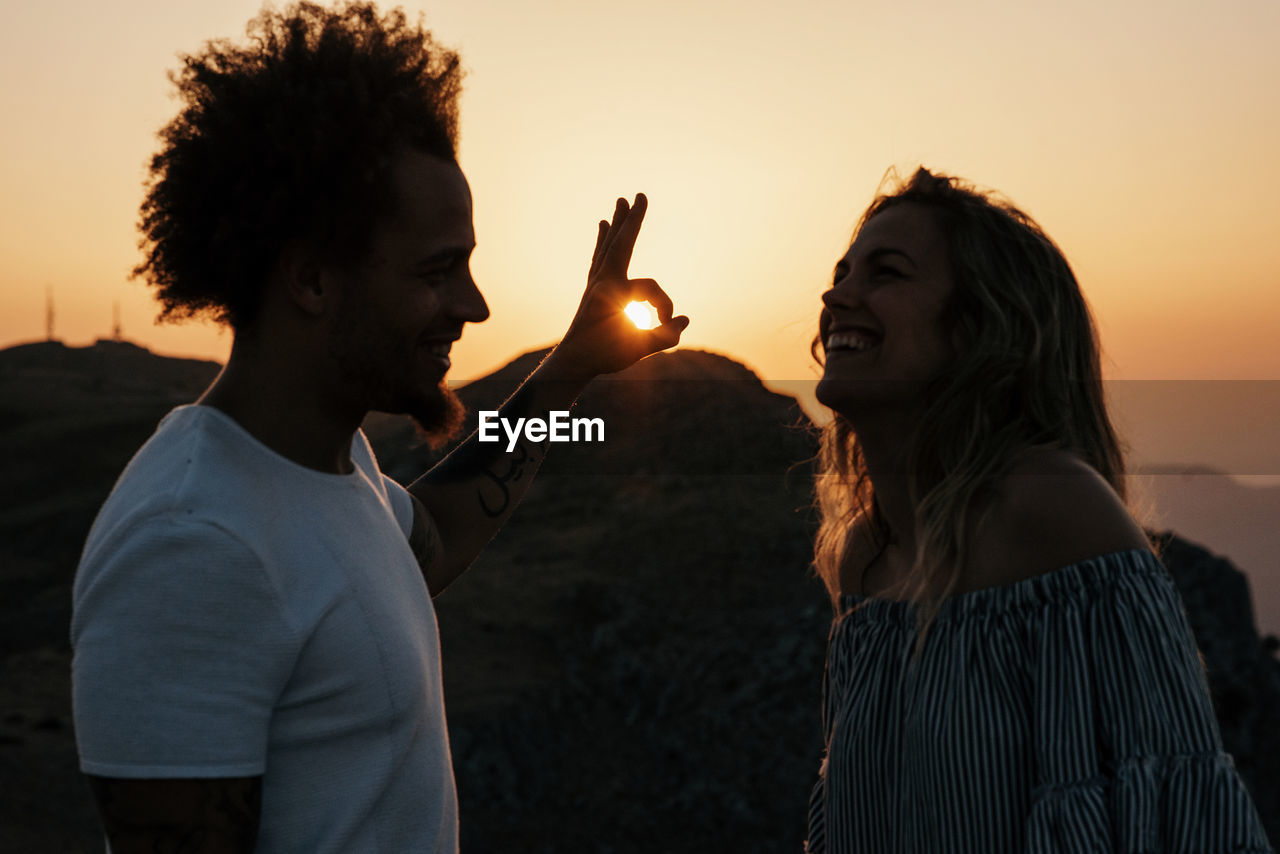 Side view of smiling hipster showing ok against sunbeams and looking at trendy woman laughing while standing against mountain ridge under colorful sky during sunset