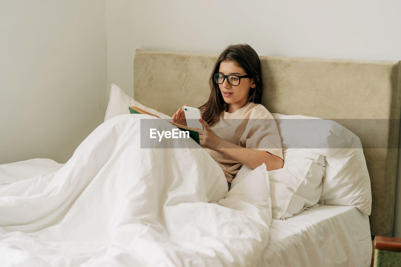 Female pretty student reading a book and taking notes on mobile phone while lying in bed