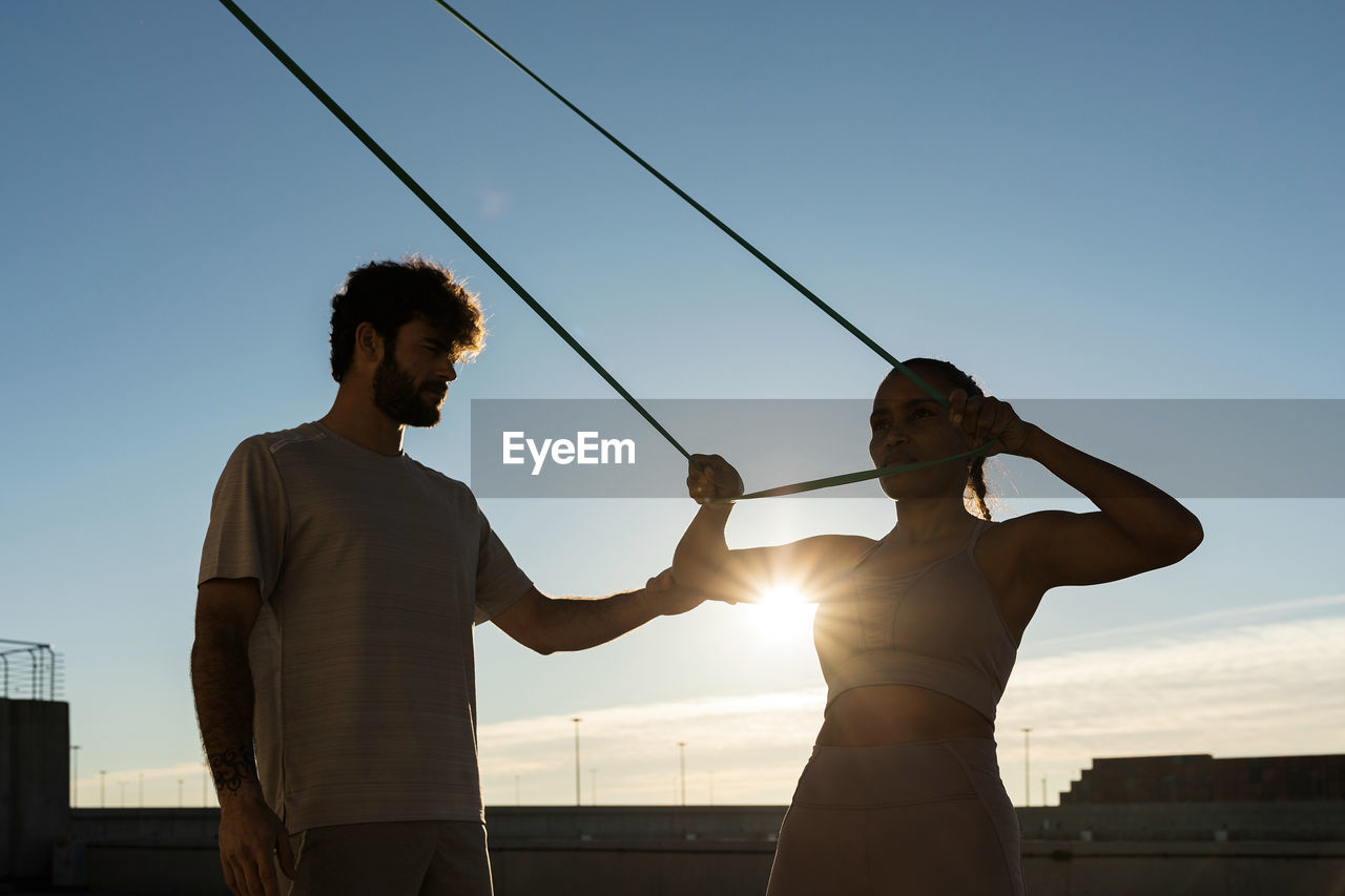 Young bearded sportsman supporting smiling ethnic female athlete during suspension training under blue cloudy sky with shiny sun in back lit