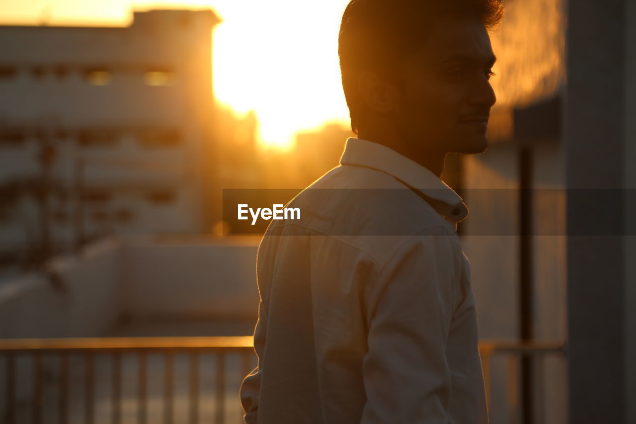 Man looking away while standing against buildings during sunset