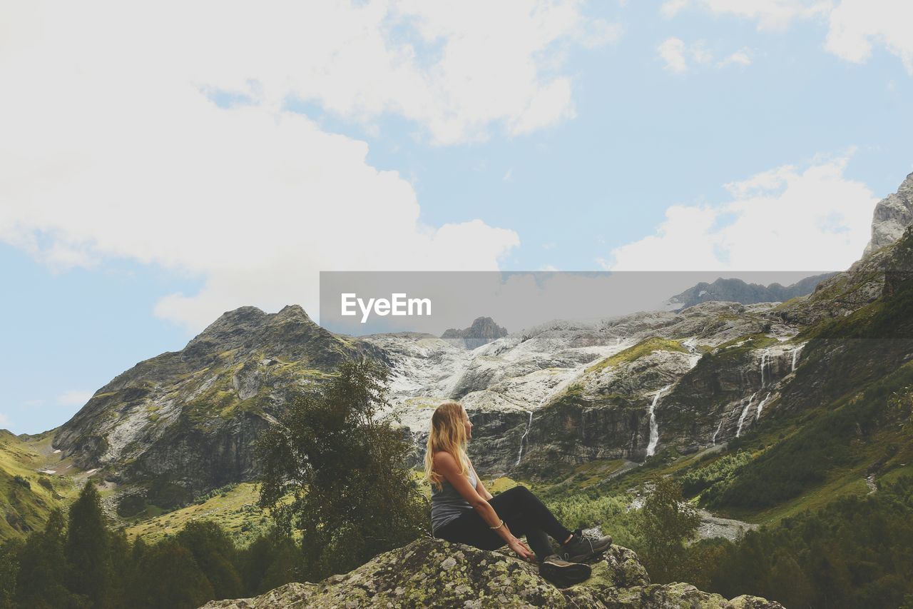 Side view of female hiker relaxing on rock at mountains against sky